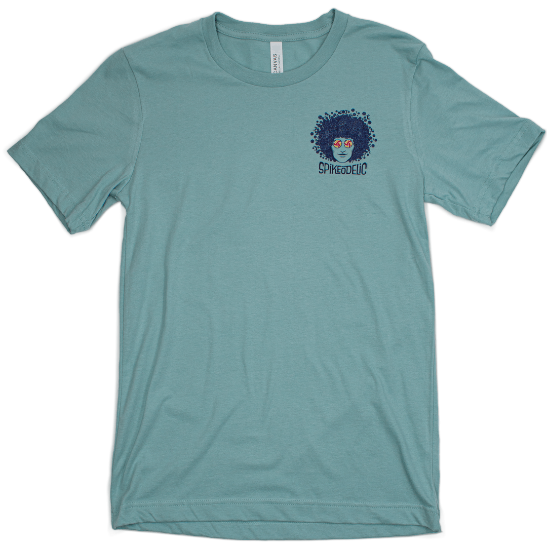 Spikeodelic volleyball men's t-shirt in dusty blue. Front pocket graphic is man's face with afro made from volleyballs and players spiking volleyballs. Man's glasses are round and feature volleyballs. The words "Spikeodelic" are below the graphic. The maj